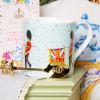 Pale blue mug decorated with guardsmen and flag. On the blue background are gold stars and on the inside of the mug is the gold coronet. A gold spoon is next to the mug and in the background is a pink box of tea and a tea cosy thumbnail