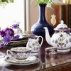Queen Victoria china tea set with a design featuring Queen Victoria's name cipher, gold plated rims and floral patterns in a palace tea table photoshoot. The set includes teapot, cream jug, pillbox and a teacup and saucer sitting on top of a side plate. thumbnail