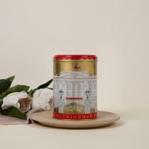 Gold tea caddy with lid off. Round teabags inside. Red lid with lion on a crown and flags surrounding. The main body of the tea caddy has the Buckingham Palace facade wrapped around. 