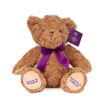 Brown teddy bear with a purple ribbon tied round its neck. Its feet are embroidered with 2022 in purple thread and on the other foot is 'Platinum Jubilee' embroidered in purple threads