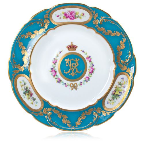 Blue and white china plate. At the centre is Queen Victoria's cypher - an entwined V & R in a blue circle with gold edge. Topped with a crown and surrounded by flowers and a bow. The edge of the plate is blue and finished with gold leaves, hand-painted fl