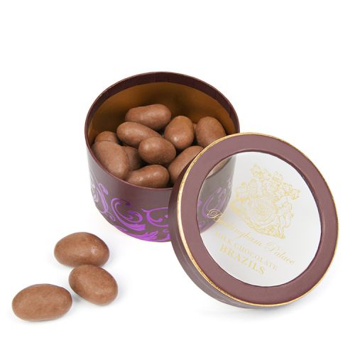 A round chocolate and purple coloured with a clear lid with gold writing stating 'Buckingham Palace Milk Chocolate Brazils'. 