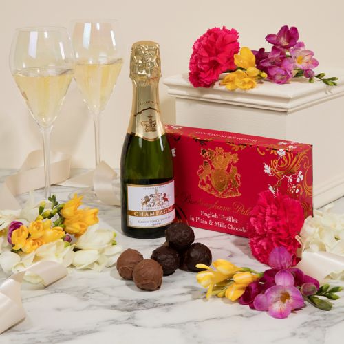 Creative image of miniature champagne bottle next to a pink box of English chocolate truffles. Next to the bottle is two glasses of champagne, some truffles and many colourful flowers