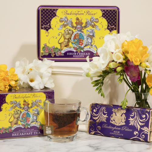 A creative shot of a purple and yellow rectangular biscuit tin on a cream plinth. In front are a yellow and purple box of breakfast tea next to a clear cup of tea and a chocolate bar on the left. White and yellow flowers are amongst the products