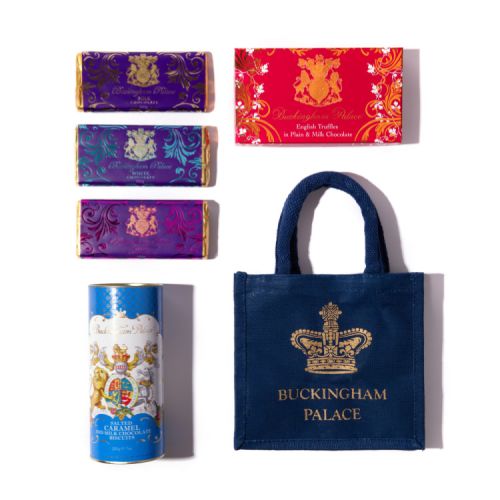 A blue gift bag with a gold crown and the words 'Buckingham Palace' on it is stood on a white cake stand. There is a gold stand of three chocolate bars in purple wrappers. A blue biscuit tube is on the left next to a pink box of chocolates.