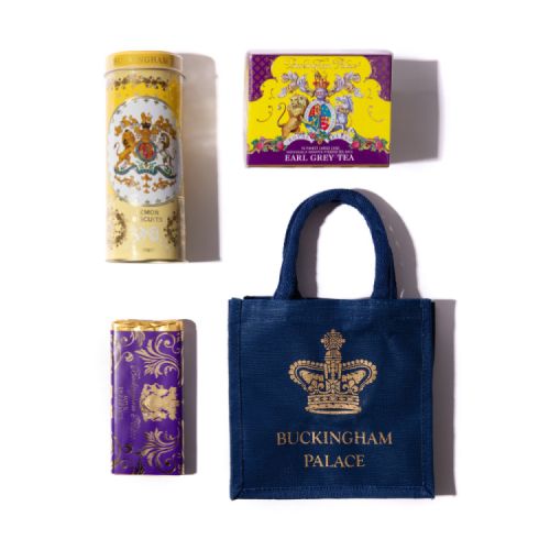 On a mahogany surface, there is a gold tray containing a yellow and purple tea box. A yellow tube of lemon biscuits is stood in a white vase of yellow flowers. There is a blue bag with a gold crown and the words 'Buckingham Palace' stood on a gold cake st