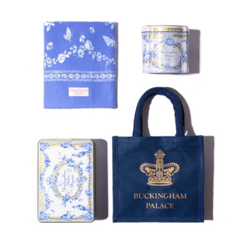 A blue bag with a gold crown and 'Buckingham Palace' is stood on a gold cake stand. Next to this is the Royal Birdsong tea caddy and biscuit tin. There is also a white vase with eucalyptus in the middle and a cup of tea on a blue tea towel
