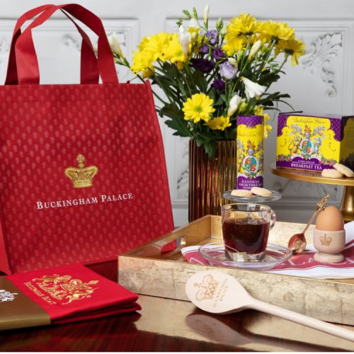 Red bag featuring a gold crown and the words 'Buckingham Palace'. It is next to a gold tray containing a cup of tea, egg in an egg cup, purple and yellow tea box and purple and yellow biscuit tube. There is also a red apron on the left and a wooden spoon.