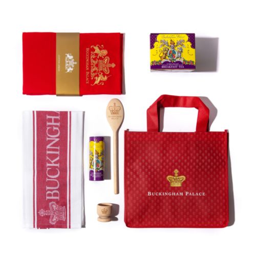 Red bag featuring a gold crown and the words 'Buckingham Palace'. It is next to a gold tray containing a cup of tea, egg in an egg cup, purple and yellow tea box and purple and yellow biscuit tube. There is also a red apron on the left and a wooden spoon.