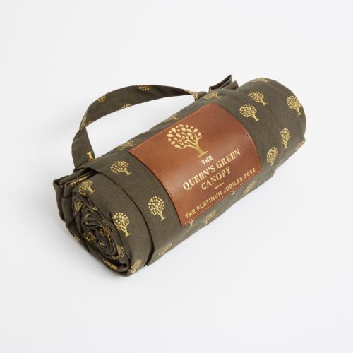 Folded up green picnic blanket printed with gold trees. Finished with a leather label printed with the words 'The Queen's Green Canopy The Platinum Jubilee 2022' and a handle