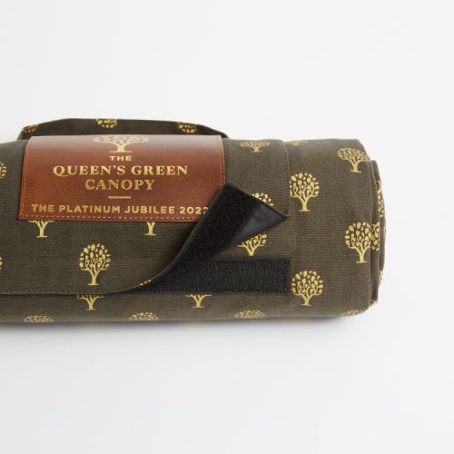 Folded up green picnic blanket printed with gold trees. Finished with a leather label printed with the words 'The Queen's Green Canopy The Platinum Jubilee 2022' and a handle