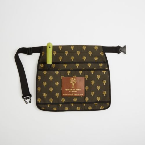 Green gardening tool belt with a black strap. The tool belt is green with gold trees printed on it and finished with a leather label printed with the words 'The Queen's Green Canopy The Platinum Jubilee 2022.' There are four pockets