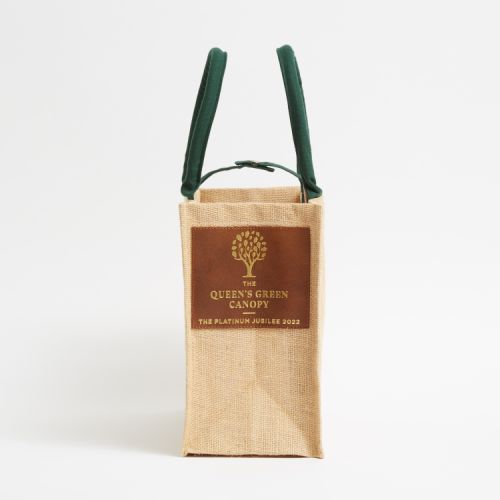 Brown juco bag printed with a green tree and the words 'The Queen's Green Canopy The Platinum Jubilee 2022.' The bag has green handles and a leather label on the side