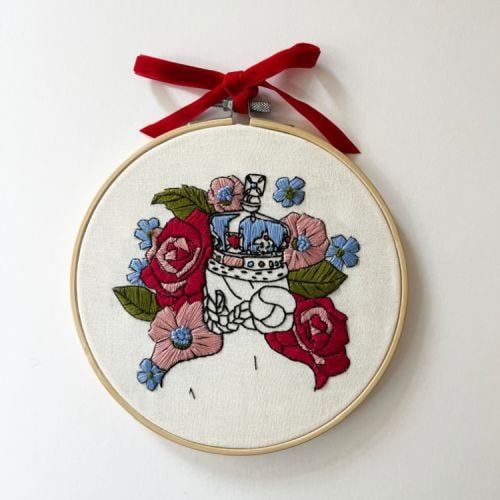 Round embroidery hoop embroidered with someone wearing a crown and surrounded by flowers and tied with a red velvet ribbon