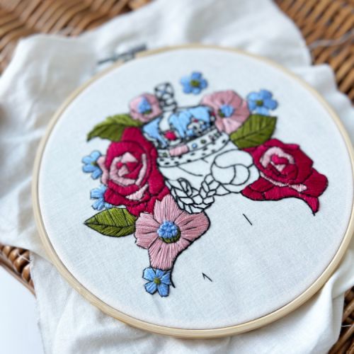 Round embroidery hoop embroidered with someone wearing a crown and surrounded by flowers and tied with a red velvet ribbon