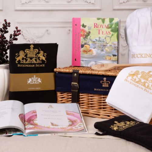 Wicker hamper with a blue lining printed with the words 'Buckingham Palace' and a gold crown. Surrounding the hamper is a black apron, oven glove and tea towel all embroidered with a gold crest. There is an open recipe book and a Royal Teas recipe book wi