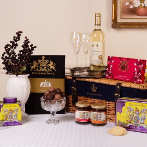 Closed wicker hamper with a pink box of truffles and a tray of two wine glasses and a bottle of white wine sat on top. In front of the hamper is a yellow and purple shortbread biscuit tin, next to a jar of strawberry jam and marmalade. On the left is a bl