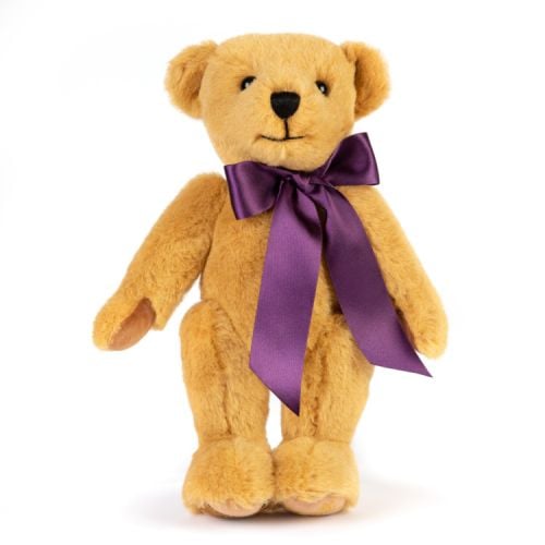 Merrythought Teddy Bear wearing a purple satin ribbon round its neck. The left foot has the Holyrood stag embroidered in gold thread. On the right foot is a label stating that the bear is exclusively made for Palace of Holyroodhouse by Merrythought