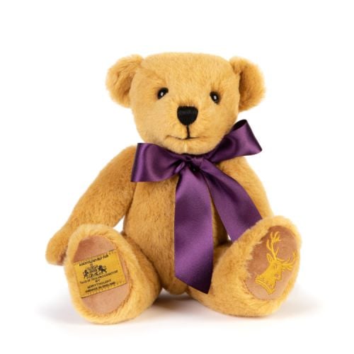 Merrythought Teddy Bear wearing a purple satin ribbon round its neck. The left foot has the Holyrood stag embroidered in gold thread. On the right foot is a label stating that the bear is exclusively made for Palace of Holyroodhouse by Merrythought