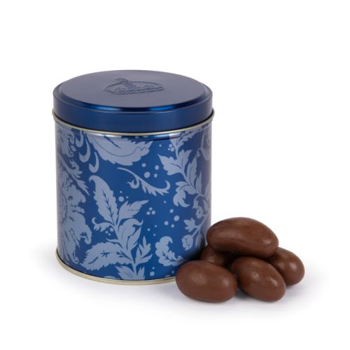 Blue tin with acanthus print and crown embellished lid. A pile of chocolate covered brazil nuts next to the tin. 