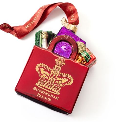 Red Buckingham Palace Christmas tree ornament shaped like a red gift box filled with colourful wrapped presents. The ornament features a gold crown emblem and the words “Buckingham Palace” on the front. A red hanging ribbon with gold “BUCKINGHAM PALACE” l