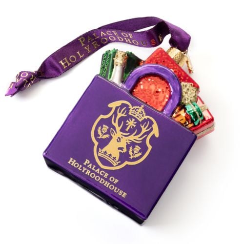 Glass Christmas ornament shaped like a purple shopping bag filled with colourful wrapped gifts. The front features the Palace of Holyroodhouse crest and gold lettering reading “PALACE OF HOLYROODHOUSE.” The ornament hangs from a purple ribbon printed with