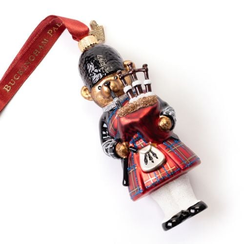 Glass Christmas ornament shaped like a teddy bear dressed as a bagpiper, wearing a red tartan kilt, black jacket and black bearskin-style hat. The bear holds red bagpipes with tartan detailing and white stockings. The ornament hangs from a red ribbon prin