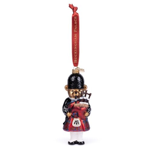 Glass Christmas ornament shaped like a teddy bear dressed as a bagpiper, wearing a red tartan kilt, black jacket and black bearskin-style hat. The bear holds red bagpipes with tartan detailing and white stockings. The ornament hangs from a red ribbon prin