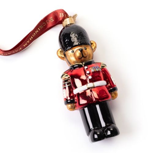 Glass Christmas ornament shaped like a King's Guard in traditional uniform, featuring a bright red tunic, black trousers and a black bearskin-style hat. The ornament hangs from a red ribbon printed with “BUCKINGHAM PALACE” in gold lettering.