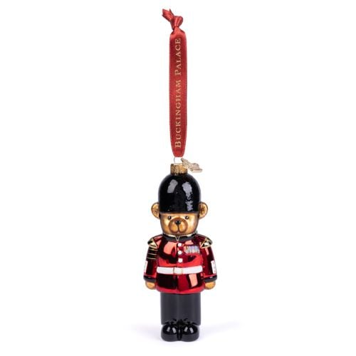 Glass Christmas ornament shaped like a King's Guard in traditional uniform, featuring a bright red tunic, black trousers and a black bearskin-style hat. The ornament hangs from a red ribbon printed with “BUCKINGHAM PALACE” in gold lettering.