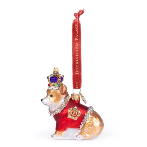 Glass Christmas ornament shaped like a corgi wearing a red ceremonial coat trimmed with sparkling gemstones and a jewel-encrusted crown. The ornament features gold decorative details and hangs from a red ribbon printed with “BUCKINGHAM PALACE” in gold let