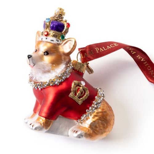 Glass Christmas ornament shaped like a corgi wearing a red ceremonial coat trimmed with sparkling gemstones and a jewel-encrusted crown. The ornament features gold decorative details and hangs from a red ribbon printed with “BUCKINGHAM PALACE” in gold let