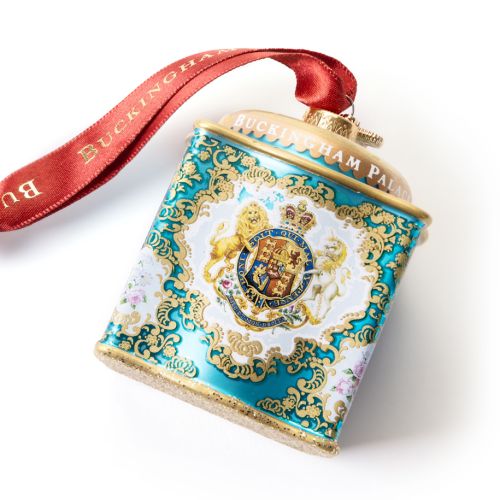 Glass Christmas ornament shaped like an ornate turquoise decorative tin, featuring the Royal Coat of Arms surrounded by intricate gold scrollwork and floral details. The lid is trimmed with gold accents and the ornament hangs from a red ribbon printed wit