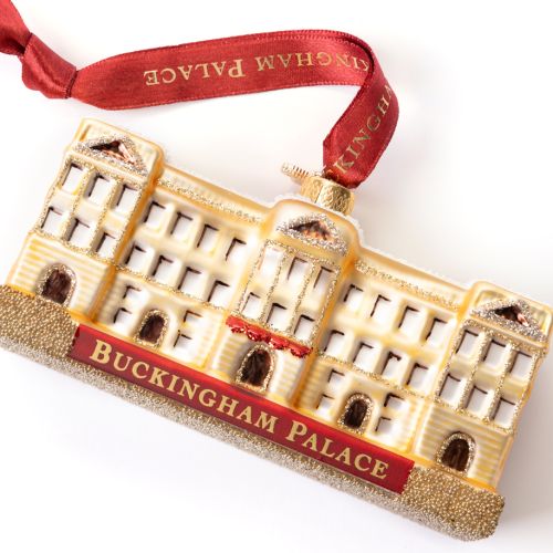 Glass Buckingham Palace ornament featuring a cream and gold representation of the palace with a snow-covered roof and glitter detailing. The front displays a red sign with gold lettering reading “Buckingham Palace.” The ornament hangs from a red ribbon pr