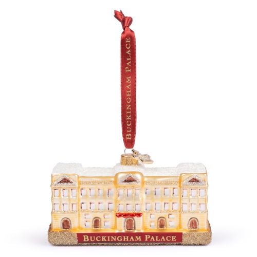 Glass Buckingham Palace ornament featuring a cream and gold representation of the palace with a snow-covered roof and glitter detailing. The front displays a red sign with gold lettering reading “Buckingham Palace.” The ornament hangs from a red ribbon pr