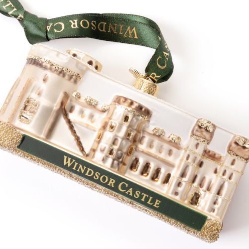 Glass Windsor Castle Christmas ornament featuring a cream and pale gold depiction of the castle with glitter accents and a snow-covered roof. The ornament sits on a glittered base with a dark green plaque reading “WINDSOR CASTLE” in gold lettering. A matc