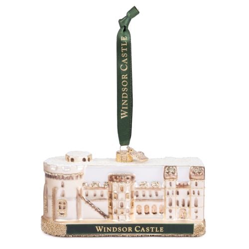 Glass Windsor Castle Christmas ornament featuring a cream and pale gold depiction of the castle with glitter accents and a snow-covered roof. The ornament sits on a glittered base with a dark green plaque reading “WINDSOR CASTLE” in gold lettering. A matc