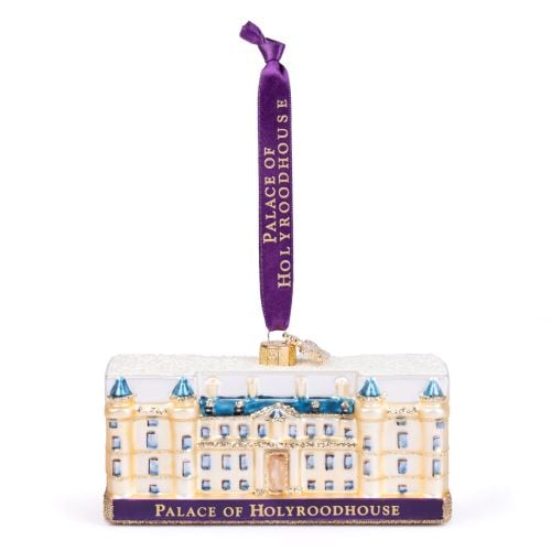 Glass Palace of Holyroodhouse Christmas ornament featuring a cream-coloured palace with blue roofs, blue window details, gold glitter accents, and a snow-covered roof. The base displays a purple plaque with gold lettering reading “PALACE OF HOLYROODHOUSE.