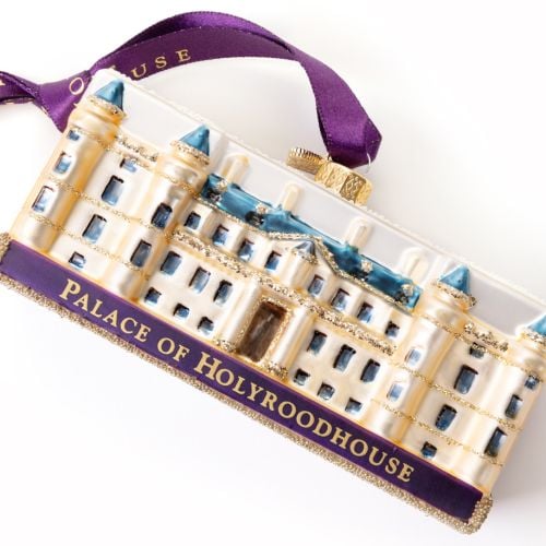 Glass Palace of Holyroodhouse Christmas ornament featuring a cream-coloured palace with blue roofs, blue window details, gold glitter accents, and a snow-covered roof. The base displays a purple plaque with gold lettering reading “PALACE OF HOLYROODHOUSE.