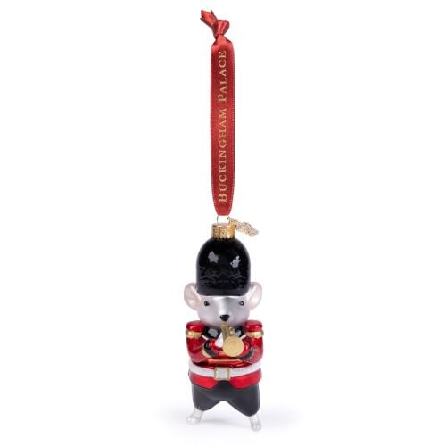 Glass Christmas ornament shaped like a mouse dressed as a ceremonial palace guard, featuring a bright red tunic with gold shoulder details, black trousers and a black bearskin-style hat. The mouse is holding a gold trumpet and hangs from a red ribbon prin
