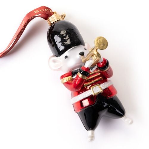 Glass Christmas ornament shaped like a mouse dressed as a ceremonial palace guard, featuring a bright red tunic with gold shoulder details, black trousers and a black bearskin-style hat. The mouse is holding a gold trumpet and hangs from a red ribbon prin