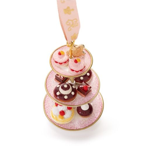 Three-tier cake stand hanging ornament featuring miniature cupcakes and iced cakes with red berry decorations on white plates. The ornament is suspended from a pale pink ribbon decorated with gold crown motifs and finished with a pink ribbon loop.