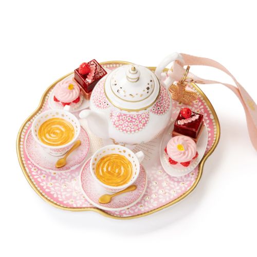 Decorative hanging ornament featuring a white and gold teapot with pink floral detailing, set on a pink serving tray with two teacups filled with tea, miniature cakes, and pink cupcakes. The ornament hangs from a pale pink ribbon decorated with gold crown