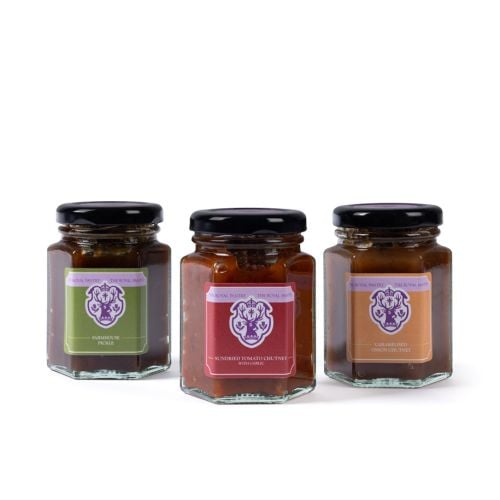 Three glass jars of The Royal Pantry condiments with black lids in a purple gift box, including Farmhouse Pickle, Sundried Tomato Chutney with Garlic, and Caramelised Onion Chutney, each featuring a distinctive coloured label and royal-inspired crest desi