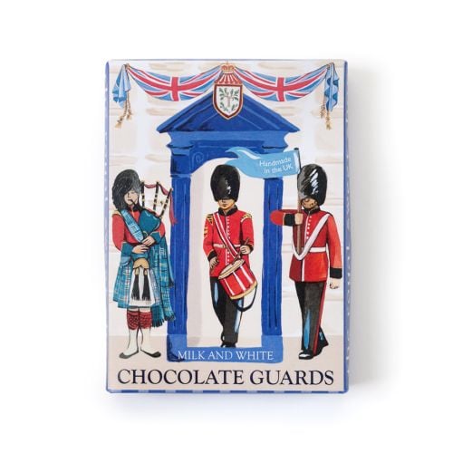 Box of “Milk and White Chocolate Guards” featuring illustrated British ceremonial guards in red uniforms and tall black hats, with a bagpiper and drummer standing beneath a blue arch decorated with Union Jack bunting. A small blue flag reads “Handmade in 