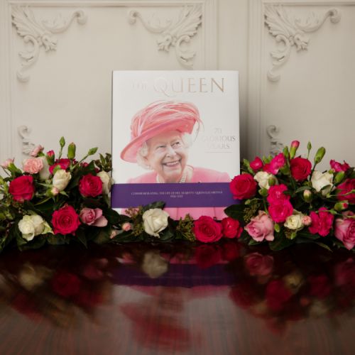 Front cover of The Queen 70 Glorious Years featuring a picture of The Queen in a pink outfit and hat. Purple band wrapped round the book to commemorate the life of Her Majesty.