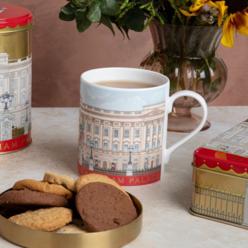 White mug with illustration of Buckingham Palace wrapping around the entire mug. Red strip at bottom with gold lettering that says 'Buckingham Palace'. Next to a matching biscuit tin and tea caddy and a gold tray of biscuits.