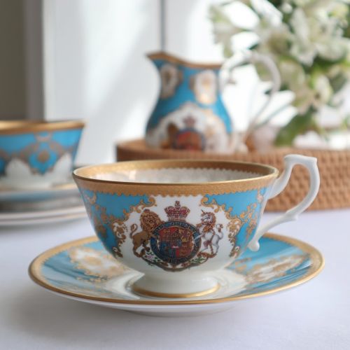Royal coat of arms fine bone china teacup and saucer featuring a gold rims and a lion and unicorn royal crest surrounded by ornated gold patterns and English flower patterns on a turquoise blue background. 