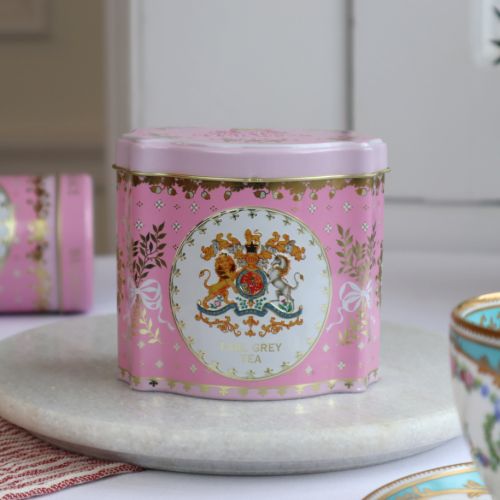 Close up shot of a pink tin tea caddy. At the centre is the Royal Coat of Arms in a white circle and edged with gold. Foliage tied with ribbon and acorns adorn the space.  