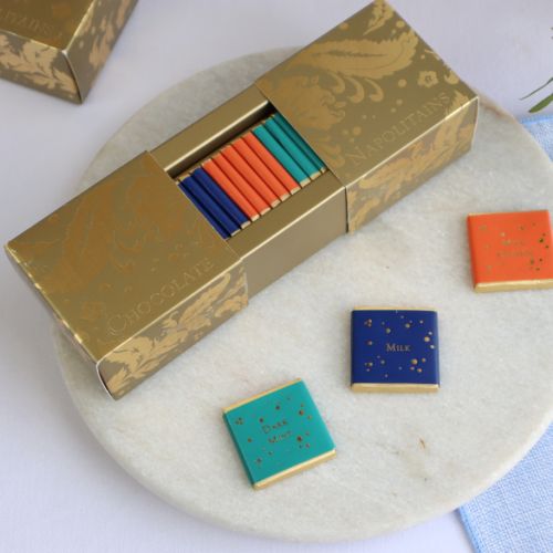 An open rectangular shaped box featuring a gold pattern, revealing at the centre, rows of individually wrapped foil chocolates with three colours and flavours, milk chocolate (dark blue), orange milk chocolate (orange) and mint dark chocolate (turquoise).